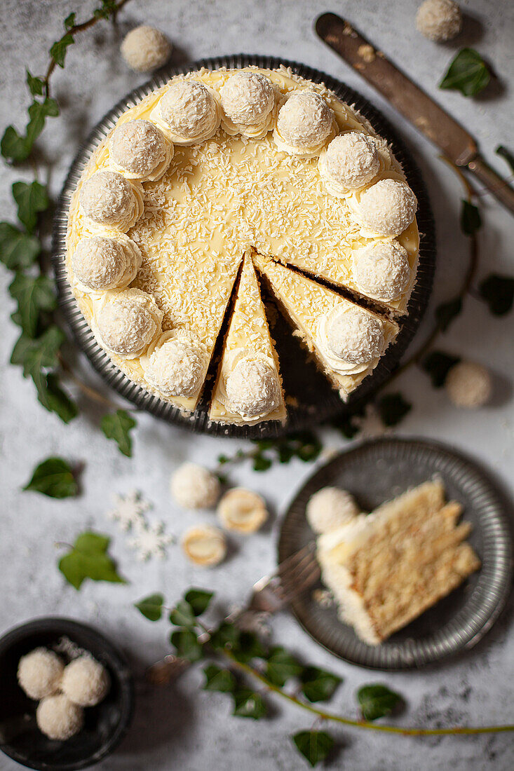 A partially sliced white chocolate and coconut cake, decorated with white chocolate ganache, cream, grated coconut and white chocolates.