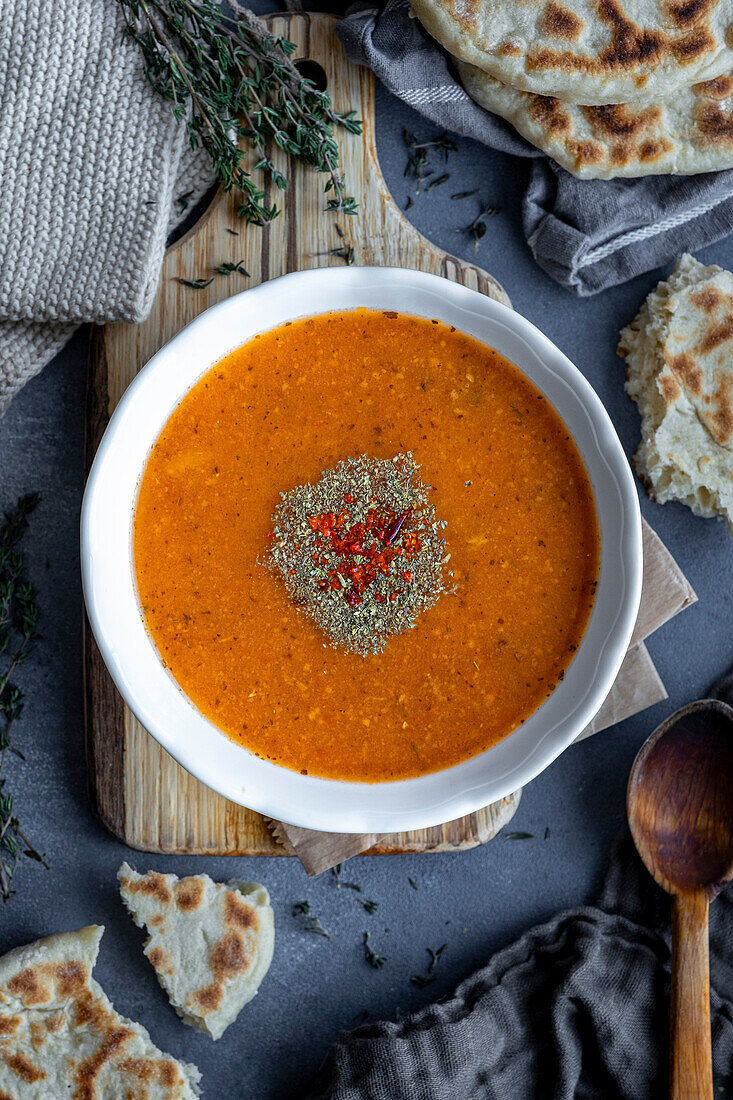 Türkische Tarhana-Suppe in einer weißen Schale, garniert mit getrockneter Minze und roten Paprikaflocken, dazu türkisches Bazlama-Brot, Thymian und ein Holzlöffel.