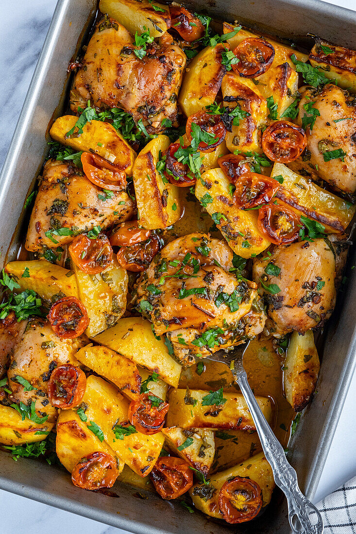 Gebackene Hähnchenschenkel, Kartoffeln und Kirschtomaten garniert mit Petersilie in einer Backform, von oben fotografiert.