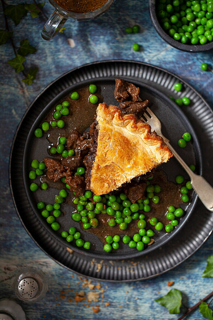 A slice of venison meat pie on a plate with peas and gravy.