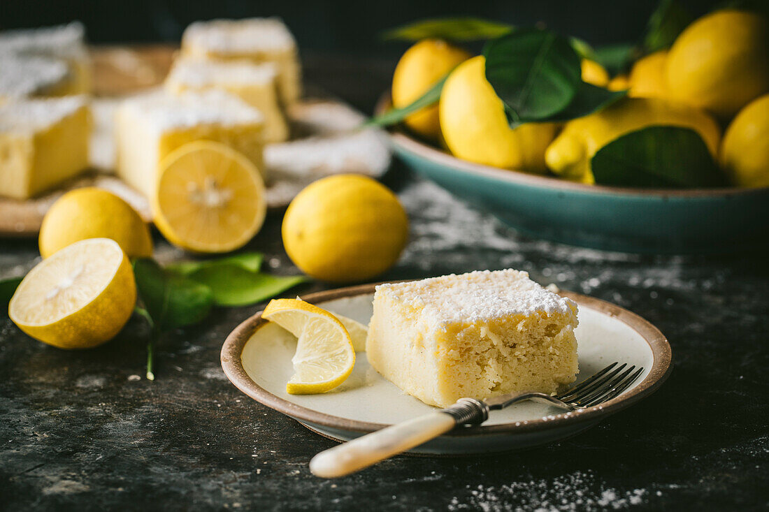 Side angle of square lemon cake slice on dessert plate, with lemon garnish and fresh lemons in background