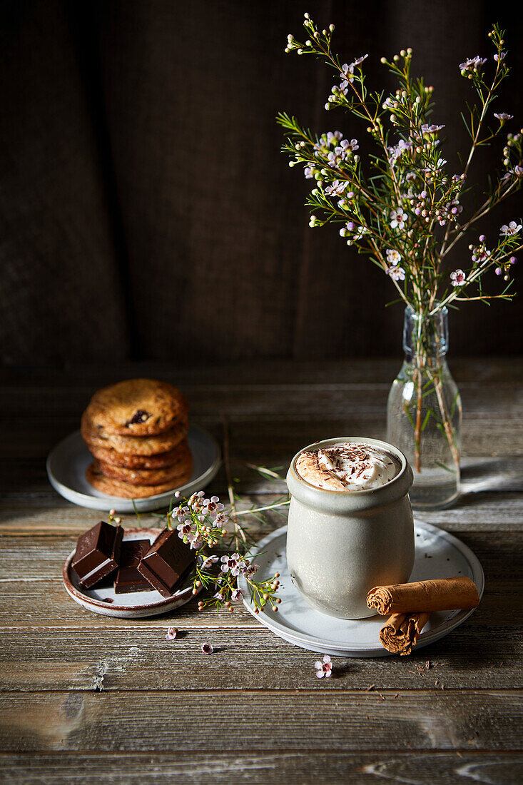 Mexican Hot Chocolate with whipped cream on a dark wood background with flowers, cookies and cinnamon sticks.