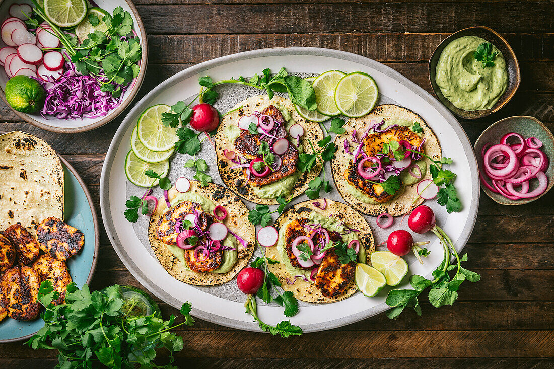 Platte mit Halloumi-Käse-Tacos mit bunten Garnierungen auf einem Tablett, mit Schalen mit Gemüse, Guacamole und eingelegten roten Zwiebeln