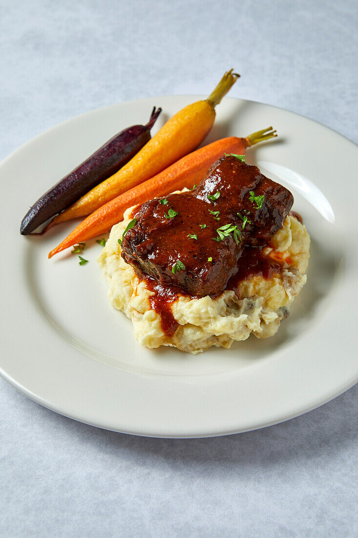 Beef short ribs with mashed potatoes and rainbow carrots on a neutral background with soft shadows