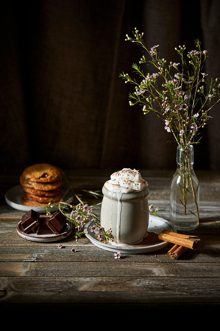 Mexikanische heiße Schokolade mit Schlagsahne auf einem dunklen Holzhintergrund mit Blumen, Keksen und Zimtstangen.