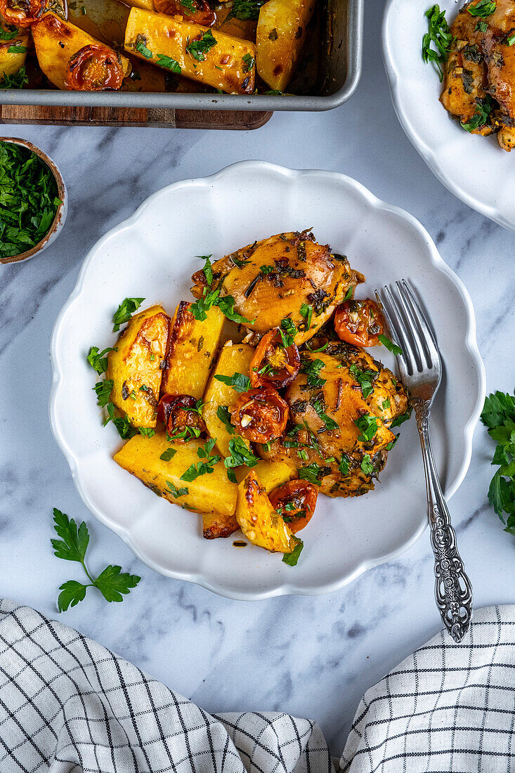 Baked chicken thighs, potatoes and cherry tomatoes, garnished with parsley in a white bowl with a fork in it, seen from above.