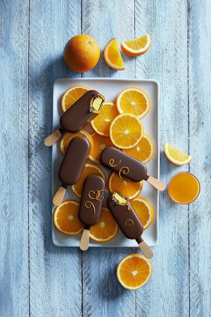 Chocolate orange popsicle ice cream on a white plate with freshly cut orange slices on a blue wooden background