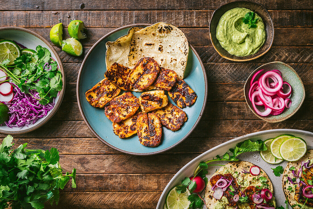 Teller mit gebratenem Halloumi-Käse, mit bunten Garnierungen in Schalen und einer Platte mit fertigen Tacos