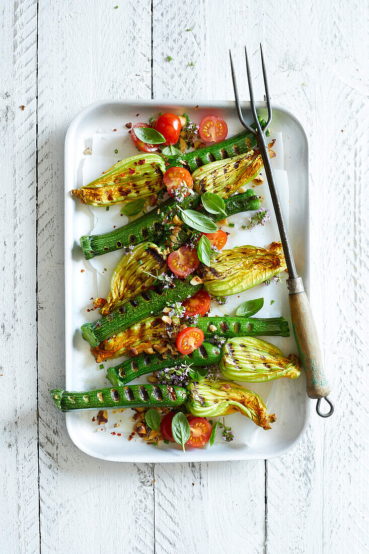 Grilled courgette flowers with cherry tomatoes, basil, chopped hazelnuts and thyme flowers on a white wooden background.