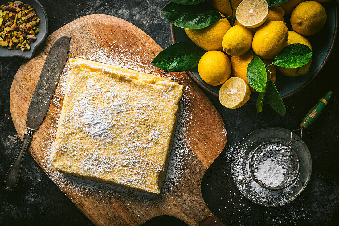 Lemon cake, unsliced, on wood cutting board with cake knife, powdered sugar, and bowl of fresh lemons