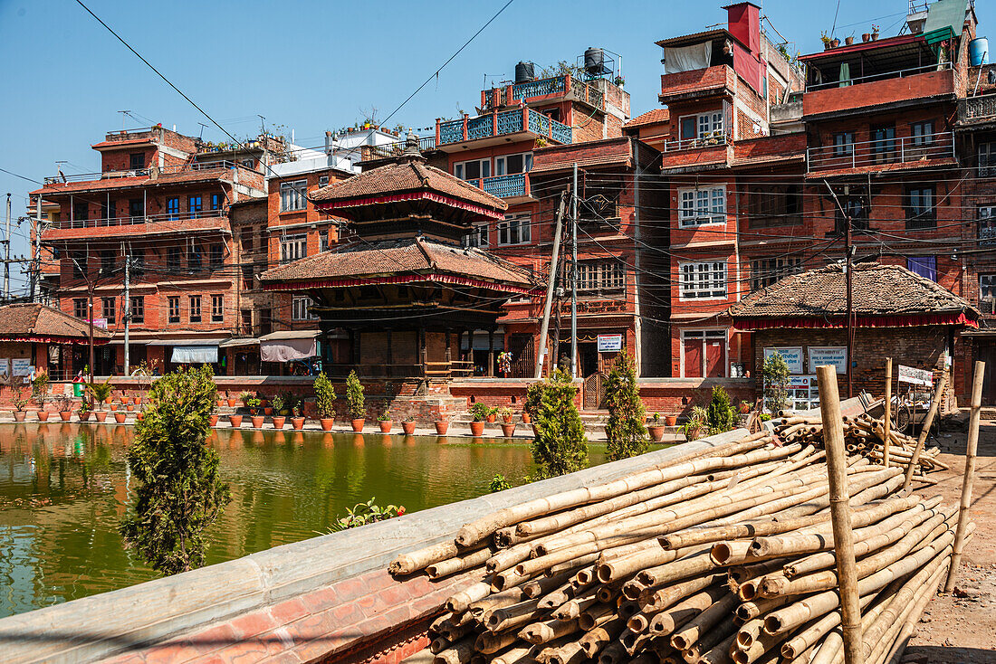 Pflanzentöpfe und Ziegelgebäude, die den See umgeben, sowie Bambusstämme, die am lebhaften grünen Bholachhe-Teich im Zentrum von Bhaktapur gelagert werden, Kathmandu-Tal, Nepal, Asien