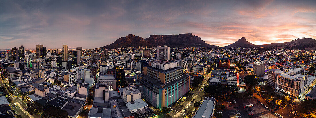 Drohnen-Panoramablick auf das Stadtbild bei Einbruch der Dämmerung, wobei die Gebäude der Innenstadt vor der Silhouette des Tafelbergs und des Löwenkopfs in einem warmen Licht erstrahlen, Kapstadt, Westkap, Südafrika, Afrika