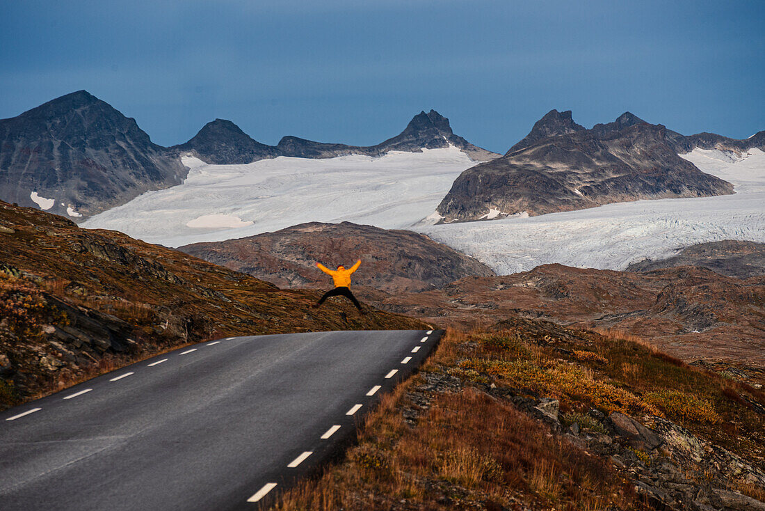 Mann in gelbem Regenmantel springt vor Gletscherbergen, Sognefjellet-Pass, Jotunheimen-Nationalpark, Norwegen, Skandinavien, Europa