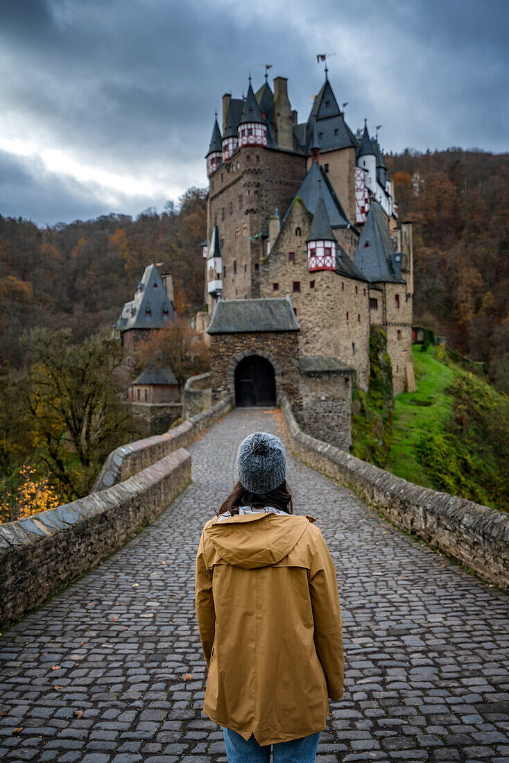 Back view of young woman with yellow jacket looking at Eltz medieval historic castle in an autumn landscape with trees at sunrise, Wierschem, Rhineland-Palatinate, Germany, Europe