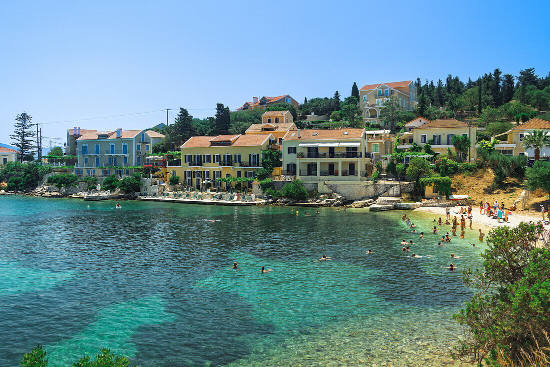 Fiscardo Dorf mit niedrigen Häusern mit roten Ziegeldächern an einem Strand mit Badenden, Fiscardo, Kefalonia, Ionische Insel, Griechische Inseln, Griechenland, Europa