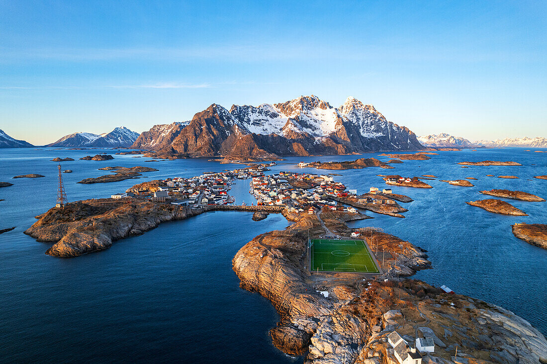 Sonnenuntergang über dem kleinen Dorf Henningsvaer, umgeben von einem Schneemassiv mit dem berühmten Fußballplatz im Vordergrund, Henningsvaer, Gemeinde Vagan, Nordland, Lofoten, Norwegen, Skandinavien, Europa