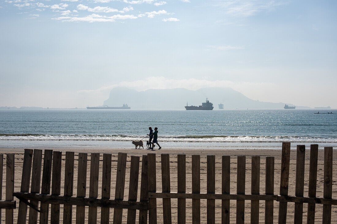 Spaziergänger mit Hund am Strand von El Rinconcillo mit Frachtschiffen in der Bucht von Algeciras und Gibraltar im Hintergrund.