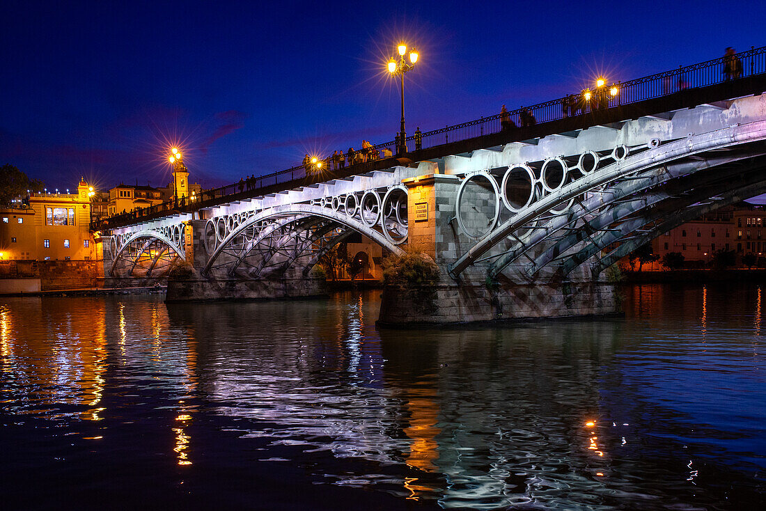 Die bei Einbruch der Dunkelheit beleuchtete Triana-Brücke mit Spiegelungen auf dem Fluss Guadalquivir in Sevilla, Andalusien, Spanien.