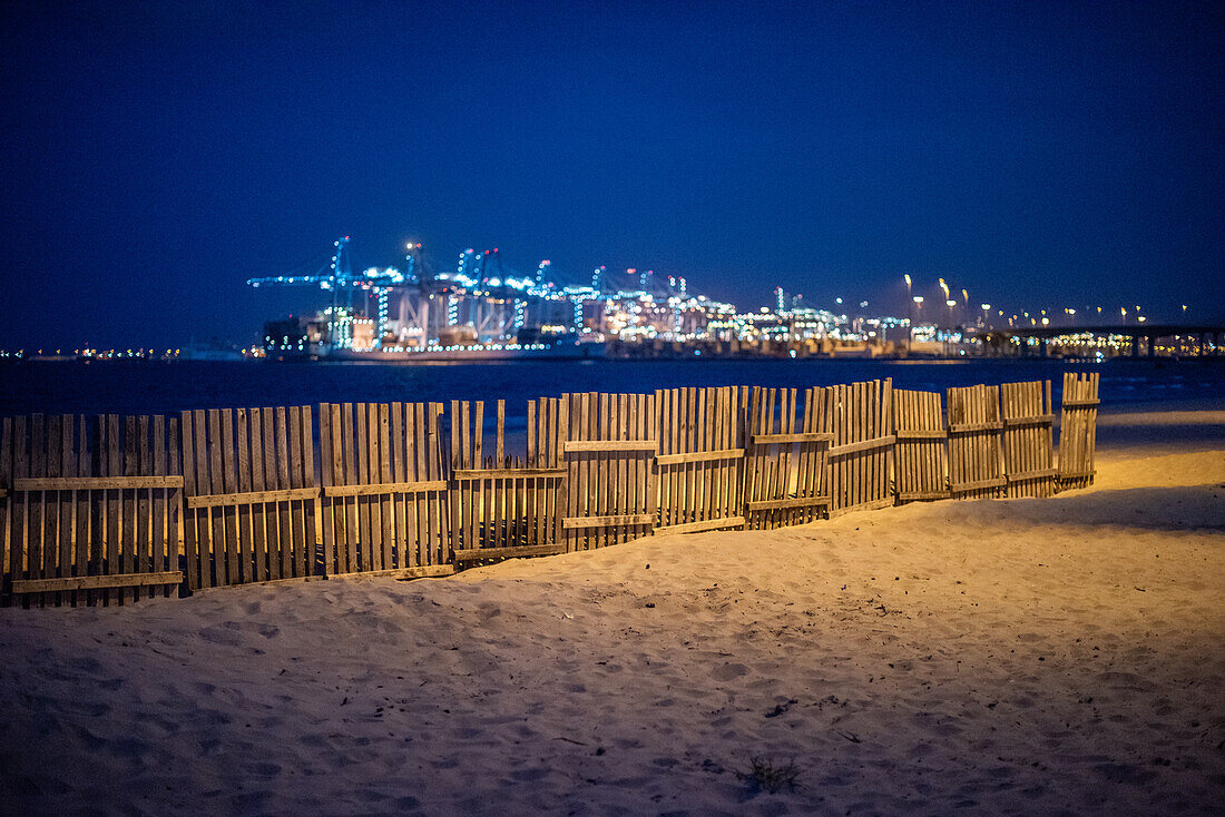 Holzzaun am Strand von El Rinconcillo mit dem beleuchteten Hafen von Algeciras im Hintergrund während der Abendzeit.