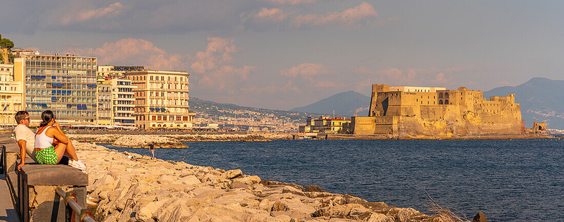 Blick auf pastellfarbene Architektur und Paar mit Blick auf das Schloss Ovo an der Uferpromenade der Via Partenope, Neapel, Kampanien, Italien, Europa