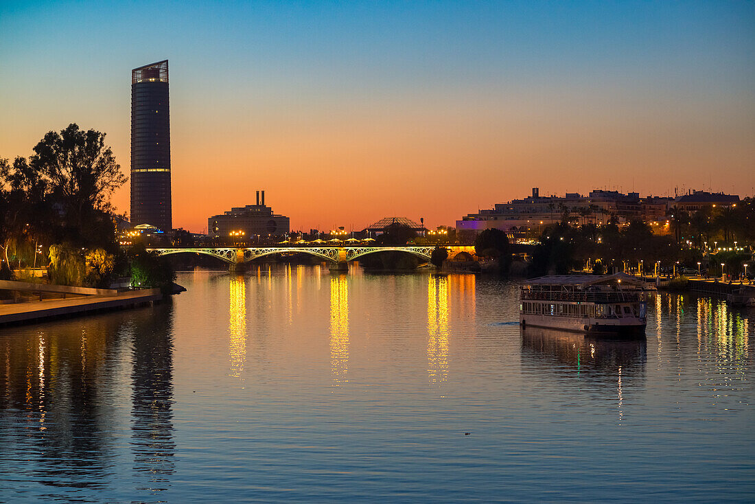 Stunning sunset view of the Guadalquivir River and the Triana Bridge in Seville, Andalucía, Spain. Evening lights reflecting on calm waters.