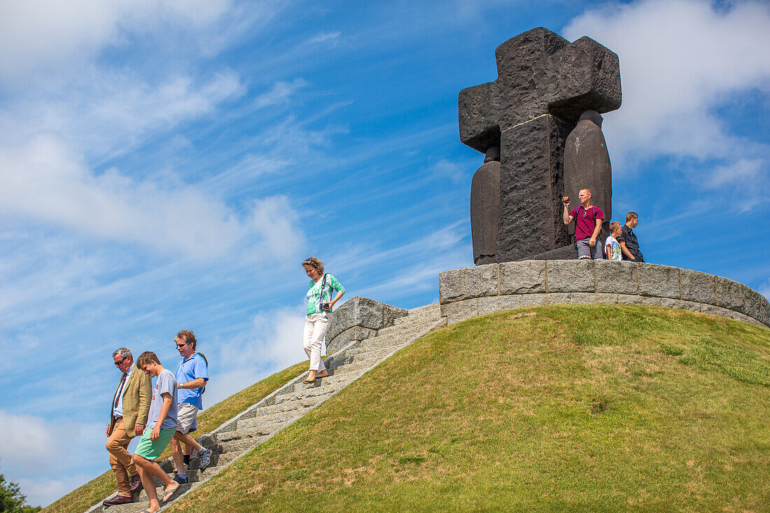 A group of visitors exploring the German military cemetery in Normandy, France, with a large cross monument in the background.