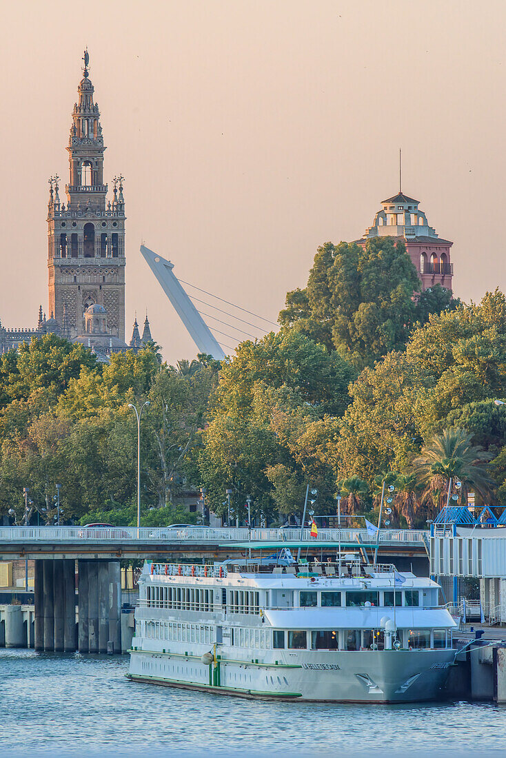 Ein Kreuzfahrtschiff angedockt am Guadalquivir-Fluss mit der Alamillo-Brücke und der Giralda im Hintergrund, Sevilla, Spanien.