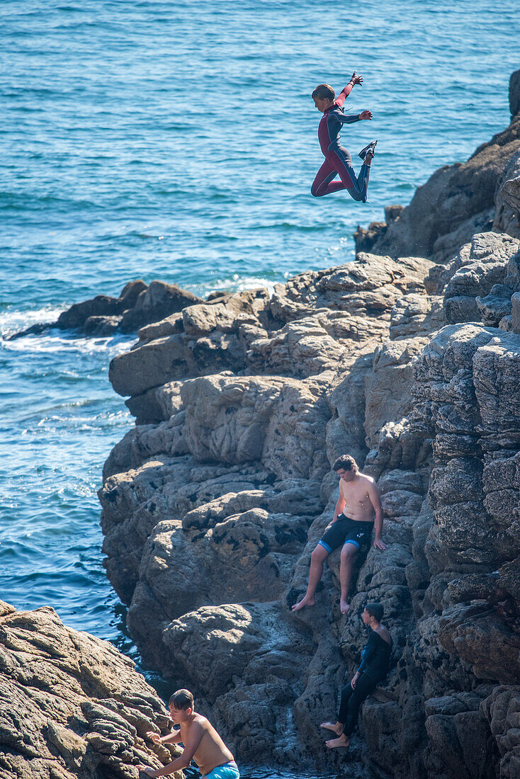 A young individual jumps off a rocky cliff into the sea in Ouistreham, Brittany, France, conveying a sense of adventure and excitement.