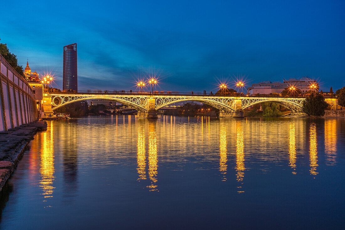 Stunning night view of the illuminated Triana Bridge reflecting on the calm waters of the Guadalquivir River in Seville, Spain.