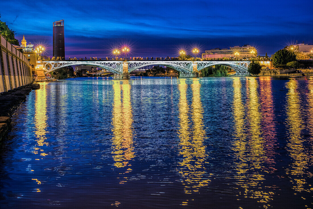 Triana Bridge illuminated at nightfall with reflections on the Guadalquivir River in Seville, Andalusia, Spain.