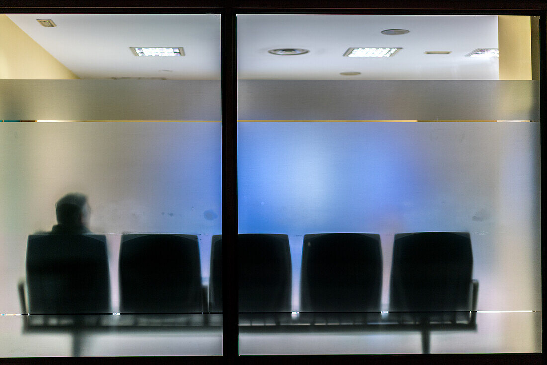 Silhouette of a man sitting alone in an empty hospital waiting room behind frosted glass, creating a sense of solitude.