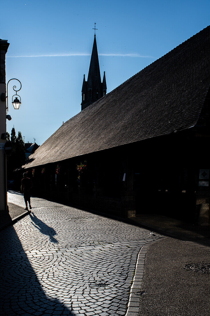 Blick bei Sonnenuntergang auf den historischen Marktplatz von Les Halles in Questembert, Bretagne, Frankreich. Mit Kopfsteinpflaster, Schatten und der Silhouette eines Kirchturms.