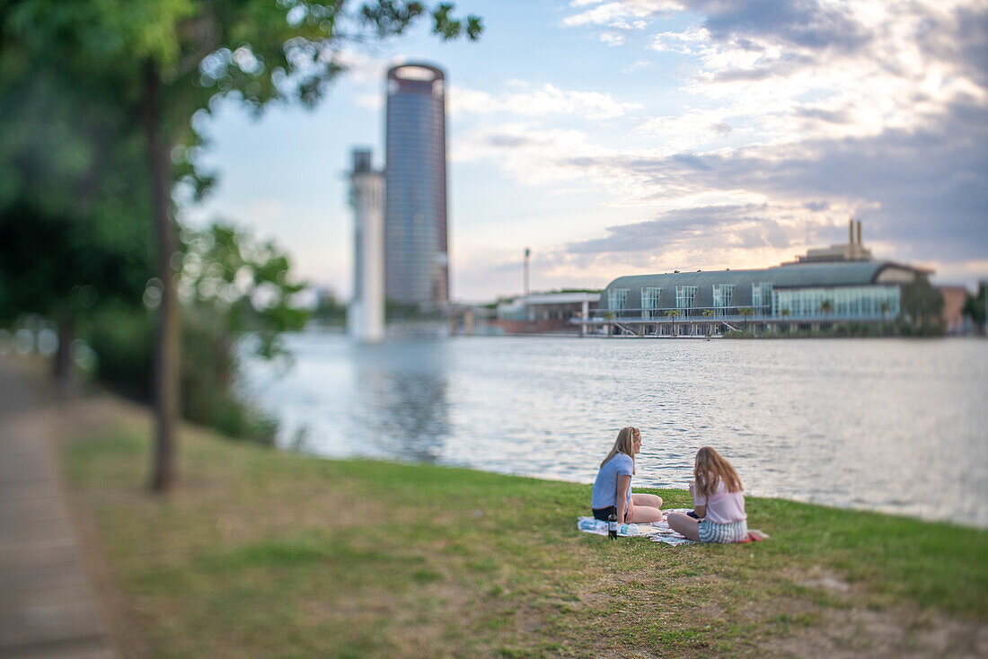 Two people enjoying a calm afternoon by the riverside in Sevilla, Spain, with a scenic view of city buildings.