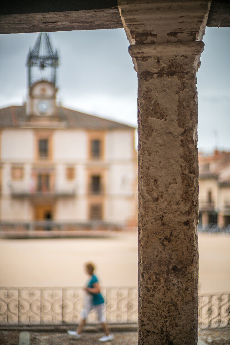 Blick auf die historische Plaza Mayor in Riaza, Segovia, Spanien, mit einer verschwommenen Figur und einem Uhrenturm im Hintergrund.