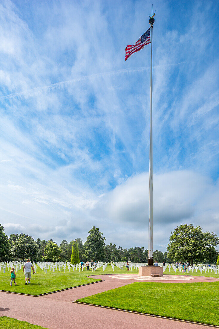 Amerikanischer Militärfriedhof in der Normandie, Frankreich, mit Reihen von weißen Kreuzen und einem hohen Fahnenmast mit der US-Flagge. Eine friedliche Umgebung zu Ehren der gefallenen Soldaten.