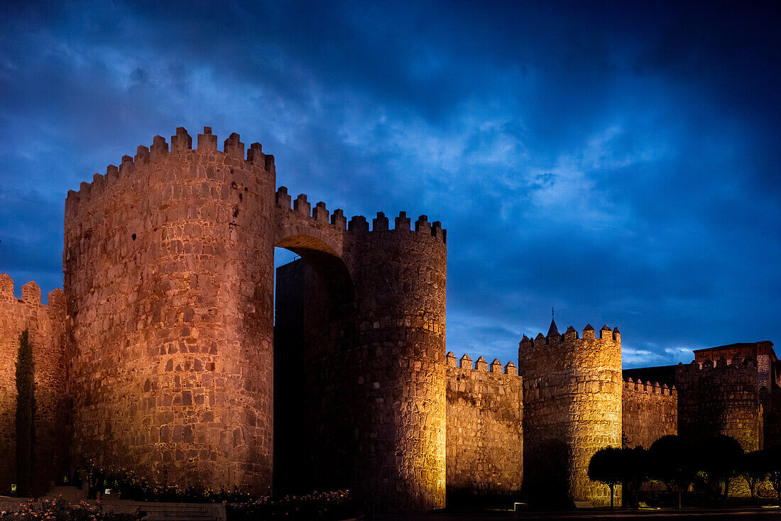 Nachtansicht der Puerta del Alcazar an der beleuchteten Stadtmauer von Avila in Castilla y Leon, Spanien. Historische Architektur vor einem dramatischen Abendhimmel.