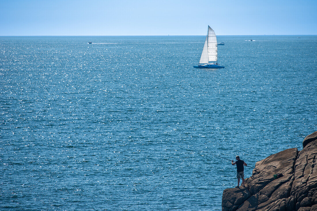 A man fishes by the rocky coast in Ouistreham, Brittany, France, with a sailing boat in the distance on a sunny day.