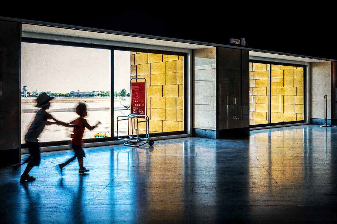 Zwei Kinder spielen und rennen im Terminal des Aeropuerto de Sevilla, Spanien. Das Sonnenlicht erhellt den Innenraum und schafft eine fröhliche und lebhafte Atmosphäre.