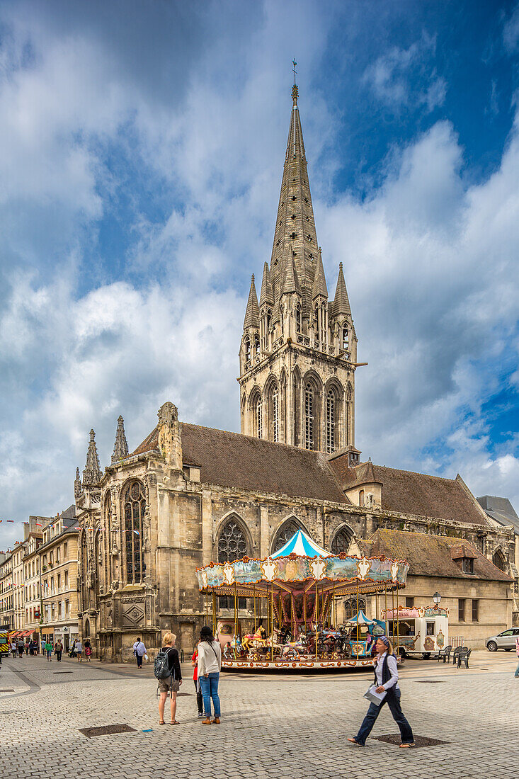 Blick auf die Eglise Saint-Sauveur in Caen, Normandie, mit einem lebhaften Karussell im Vordergrund. Die Menschen gehen spazieren und genießen den charmanten Platz.