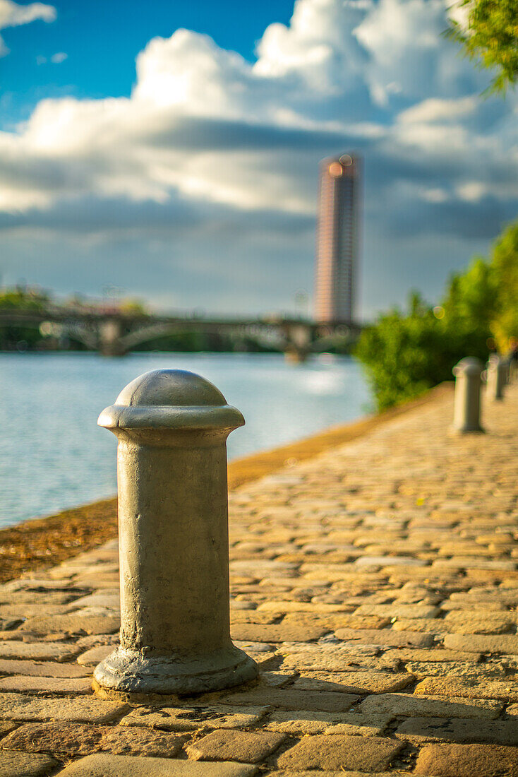 Blick auf die Uferpromenade von Sevilla, Spanien, mit Kopfsteinpflaster, Metallpfosten und dem Torre Sevilla im Hintergrund an einem bewölkten Tag.