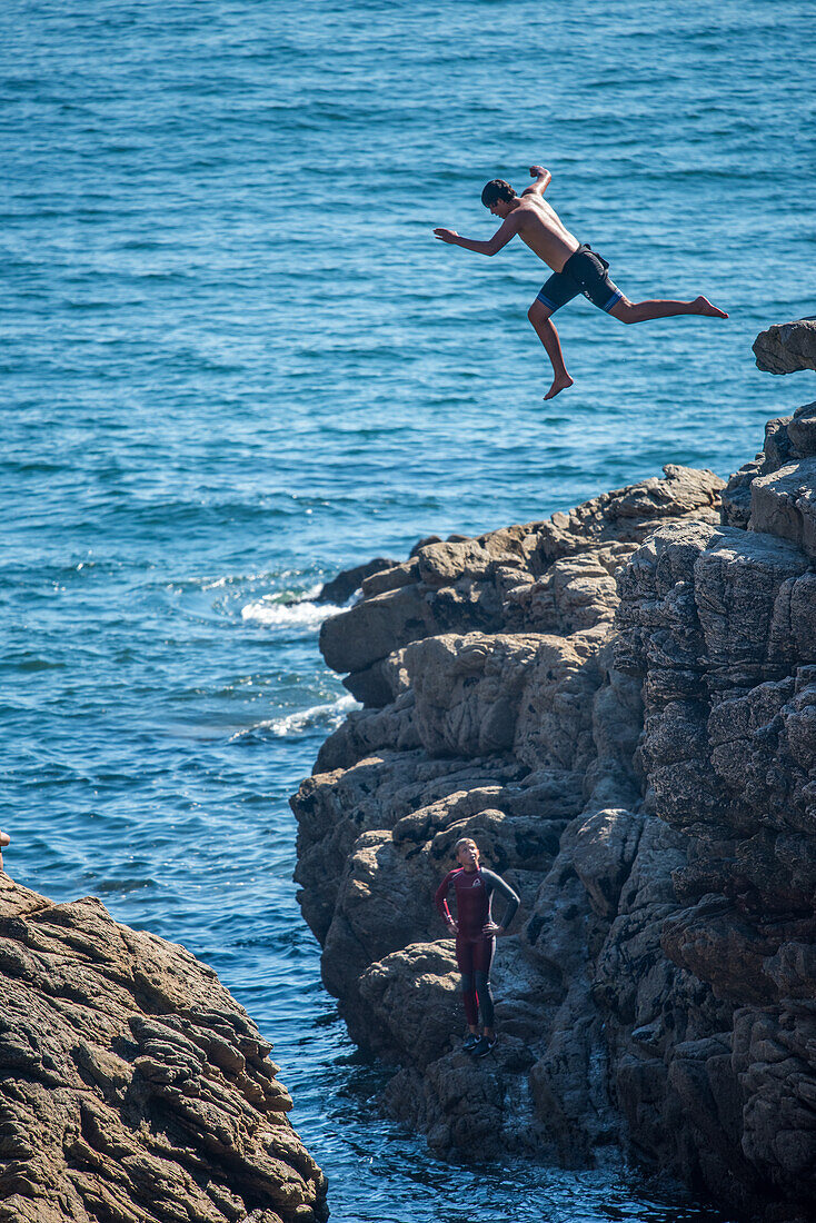 A young individual jumps off a rocky cliff into the sea in Ouistreham, Brittany, France, conveying a sense of adventure and excitement.