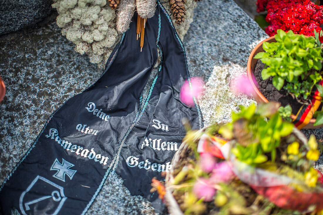 Nahaufnahme eines Waffen-SS-Gedenkbands auf dem deutschen Soldatenfriedhof in der Normandie, Frankreich, umgeben von bunten Blumen und steinernen Artefakten.