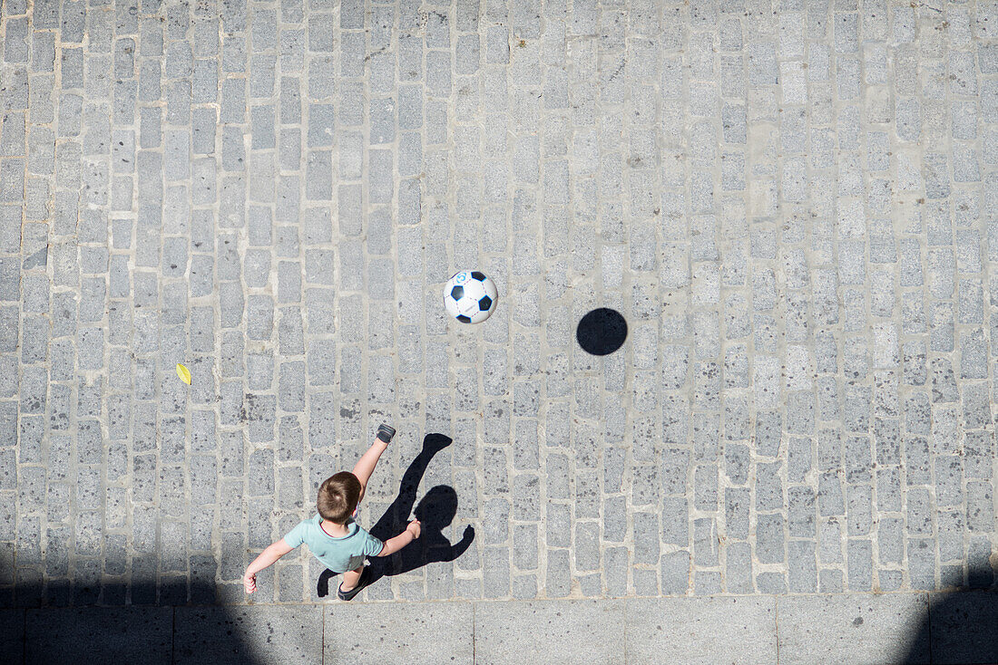 Ein kleiner Junge spielt an einem sonnigen Tag auf einer Kopfsteinpflasterstraße in Sevilla, Spanien, Fußball.