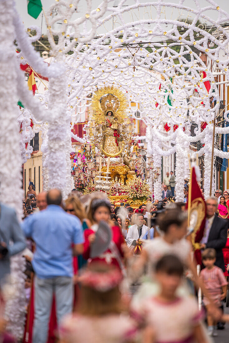 Katholische Gläubige tragen die Statue der Virgen del Rosario unter dekorativen Bögen während einer religiösen Prozession in Carrión de los Céspedes, Sevilla, Spanien.