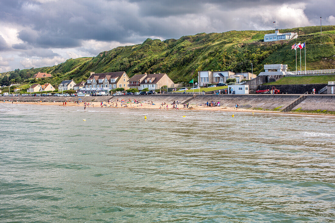 A scenic view of Omaha Beach in Normandy, France, with people enjoying the coastline and historical landmarks on a partly cloudy day.