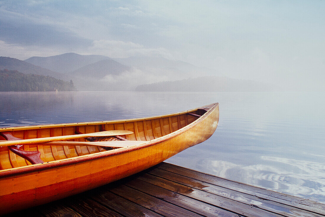 Wooden canoe on dock by calm Lake Placid covered with mist