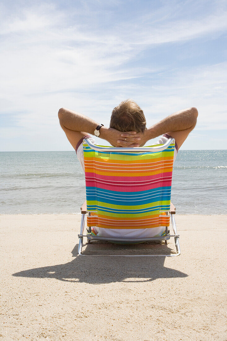 Rückansicht eines Mannes, der sich auf einem Liegestuhl am Sandstrand entspannt