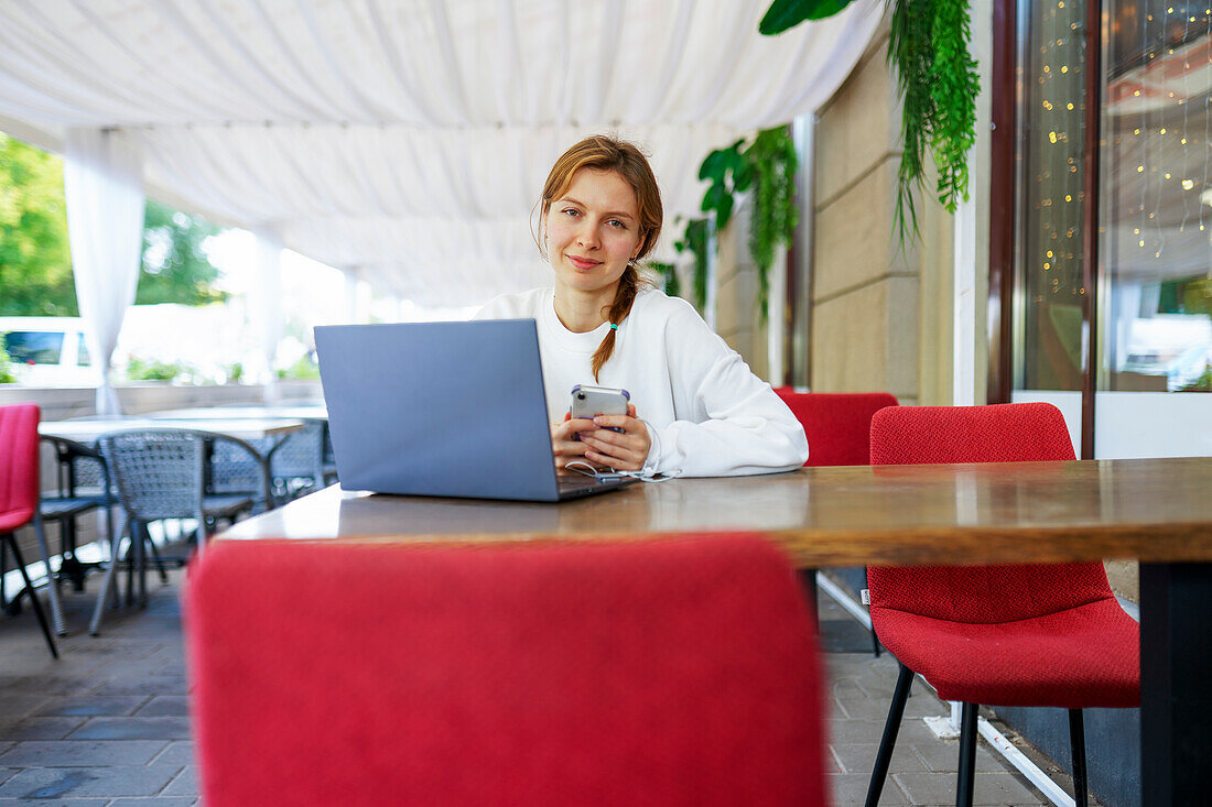 Porträt einer lächelnden Frau mit Smartphone und Laptop an einem Cafétisch