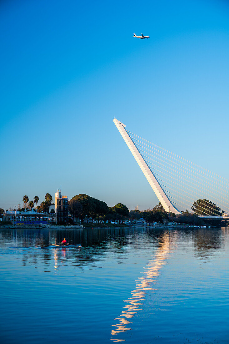 Stunning view of Puente del Alamillo in Sevilla, Andalucia, with an airplane flying overhead and calm waters reflecting the bridge.