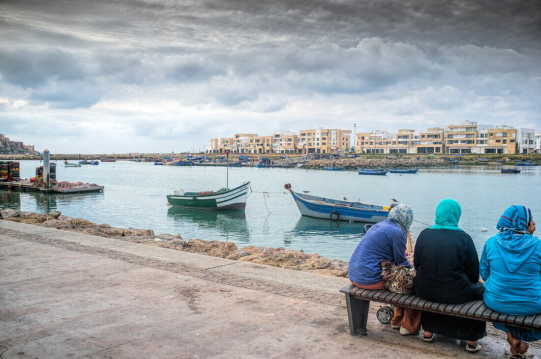 People relax along the Bou Regreg River in Rabat, enjoying the view of boats and the city under a dramatic overcast sky.